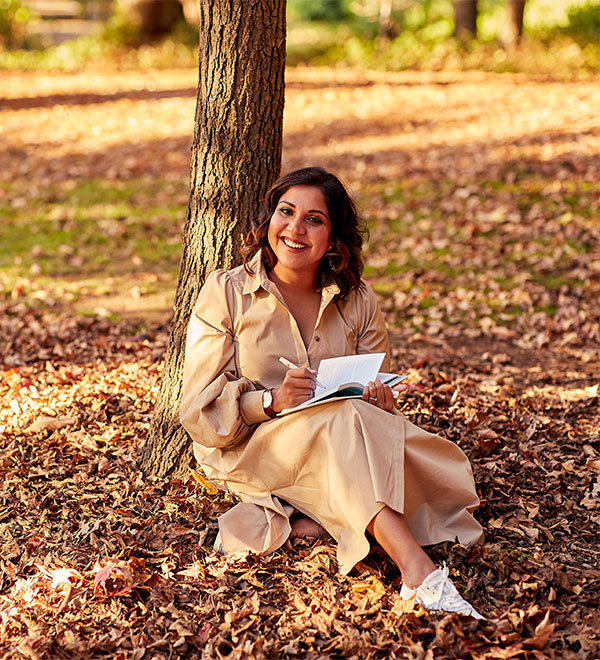 Debanjali sits under a tree in a park with a book, smiling. There are fallen leaves all around her